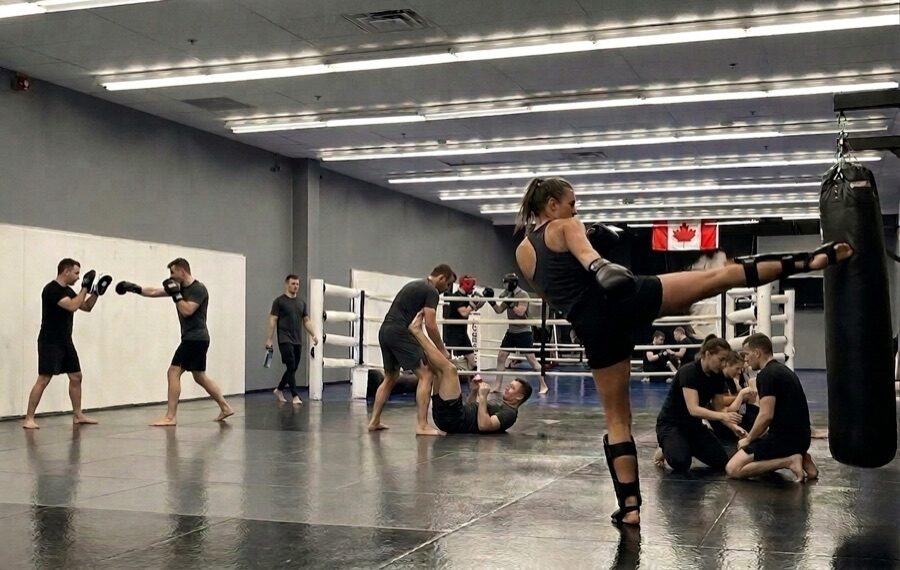 A busy adult Muay Thai class at Rumble Martial Arts — pairs on pads, a kick going into the heavy bag, the white boxing ring behind.