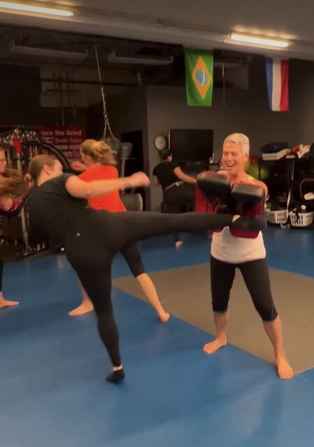 A Battle Girl member throwing a high round kick into Thai pads held by a smiling partner.