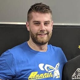 Coach Brett Milwain smiling in a blue Rumble Martial Arts t-shirt at the gym.