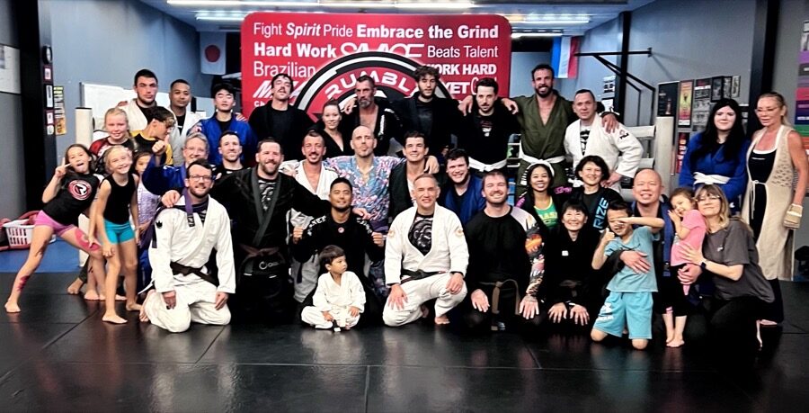 The whole Rumble gym on the mats together — kids, teens, parents and coaches in gis and rash guards, in front of the Rumble banner.