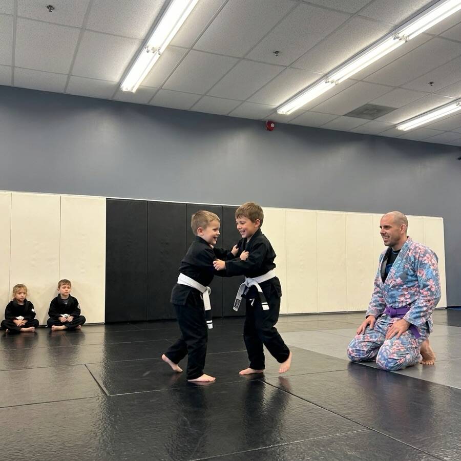 Two small children in black gis grip-fighting on the mats while a coach in a floral gi kneels beside them